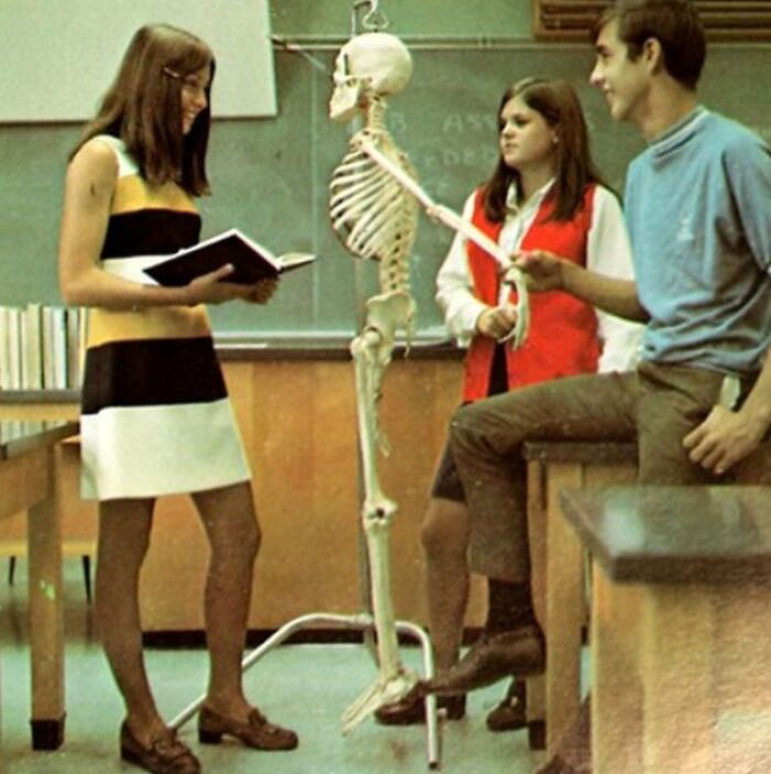 Students in a 1970s high school classroom, studying a skeleton model and reading from a book.