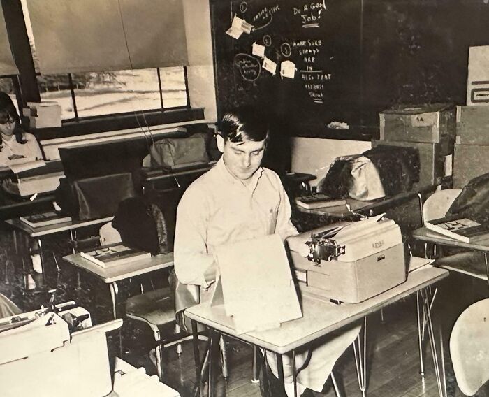 A '70s high school classroom with students using typewriters, chalkboard in background.