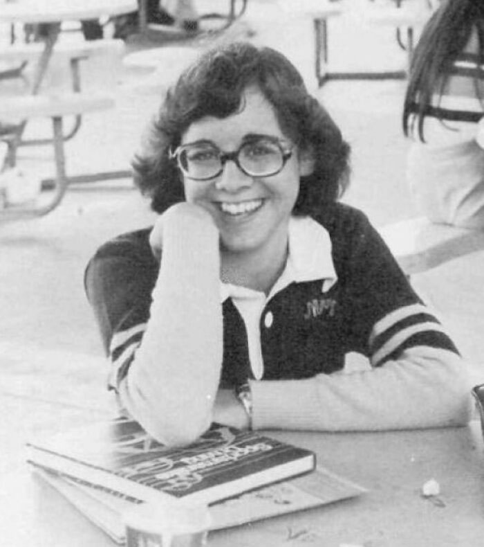 A 1970s high school student wearing glasses, smiling and sitting at a table with books.