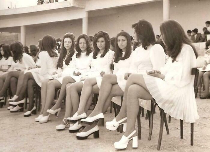 High school girls in '70s fashion, wearing white dresses and platform shoes, sitting outdoors in rows of chairs.