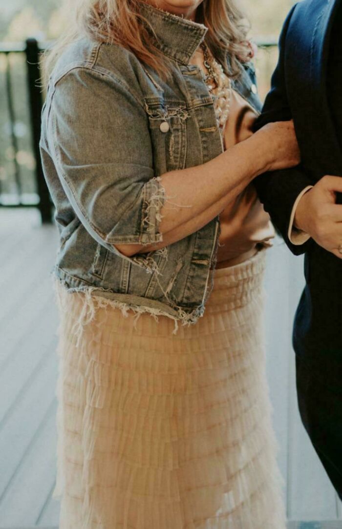 Woman at wedding wearing a frayed denim jacket over a peach ruffled dress, arm linked with a suited individual.