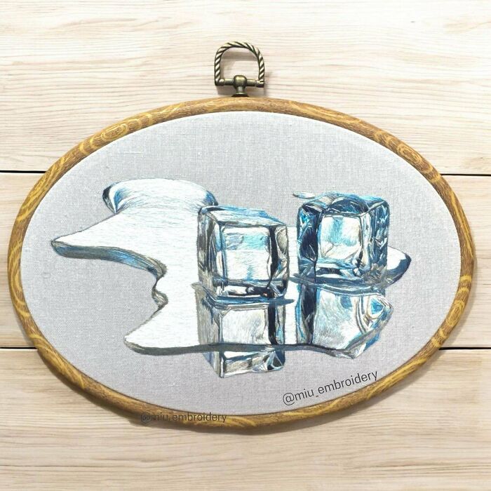 Realistic embroidery design of melting ice cubes with detailed reflections in a wooden hoop on a light wooden surface.