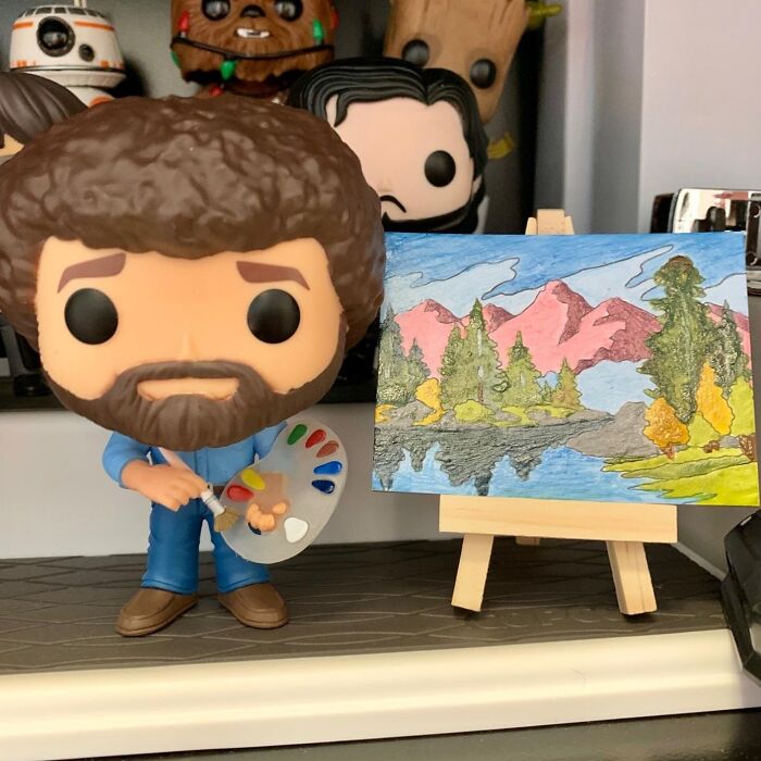 Bob Ross Funko Pop figure holding a paint palette next to a colorful landscape painting on a small easel, ideal white elephant presents.