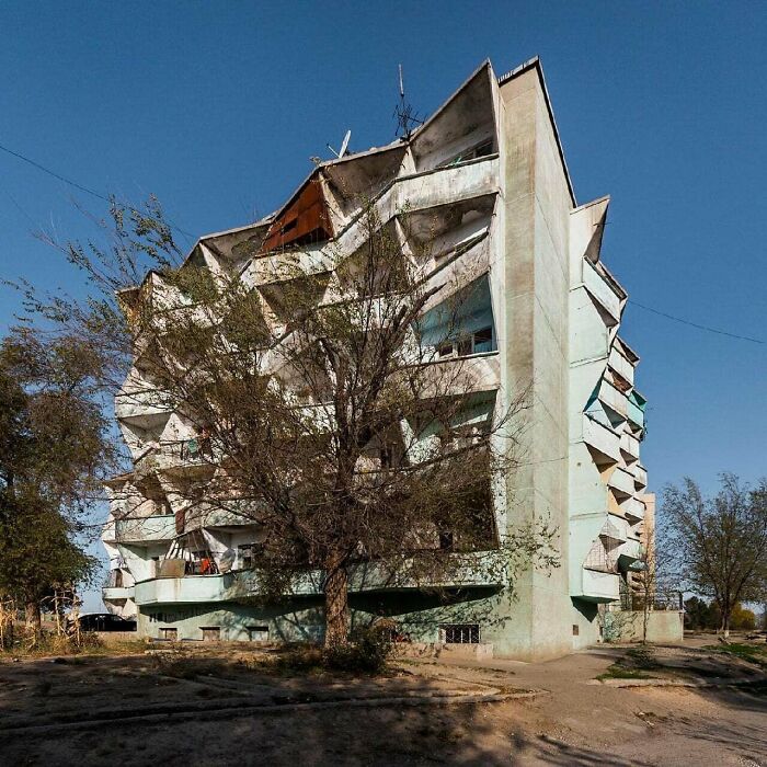 Dilapidated urban building with damaged balconies and overgrown trees under a clear blue sky, showcasing urban hell.