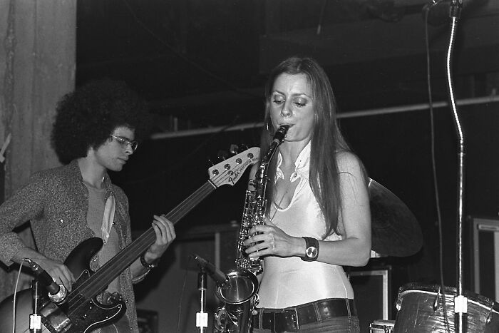 1973 musicians performing; woman playing saxophone, man with a bass guitar, capturing vintage concert vibe.
