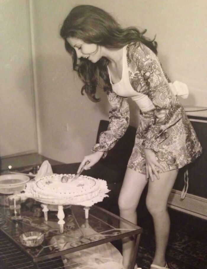 Woman in 1973 fashion cutting a cake in a living room.