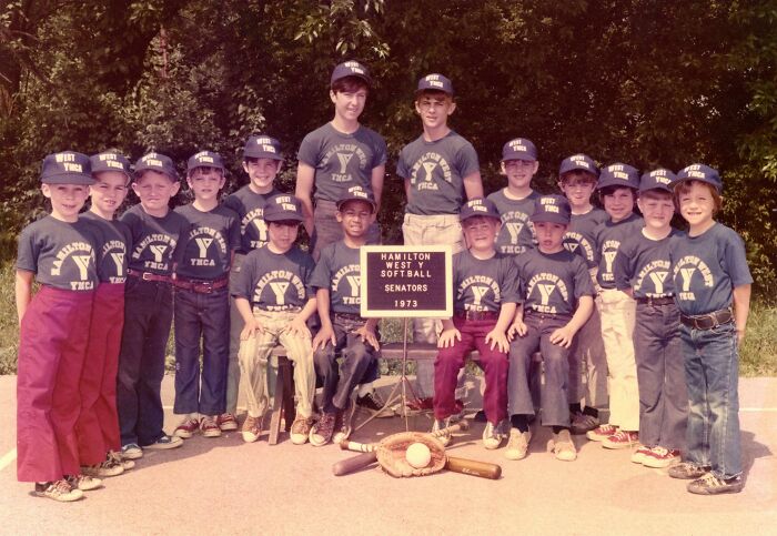 A children's softball team in 1973, wearing matching uniforms, posing with a sign and equipment outdoors.