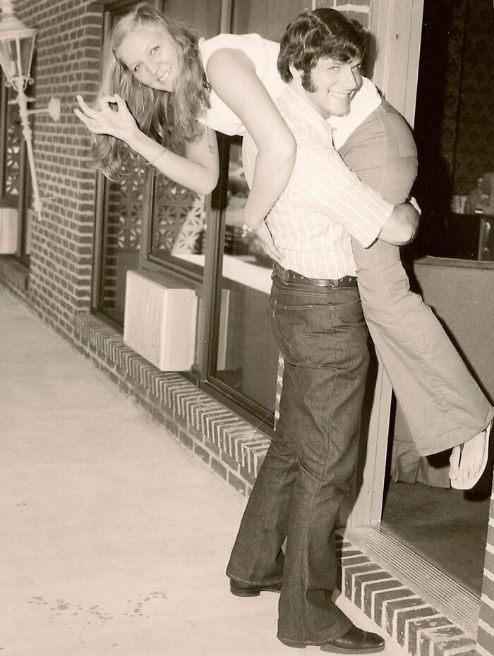 1973 retro scene: Young man carrying woman playfully indoors, both smiling warmly.