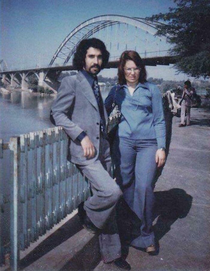A couple in 1973 posing by a bridge, both in vintage attire.