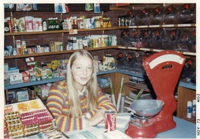 A woman in a 1973 store with shelves of products and an old-style red scale.