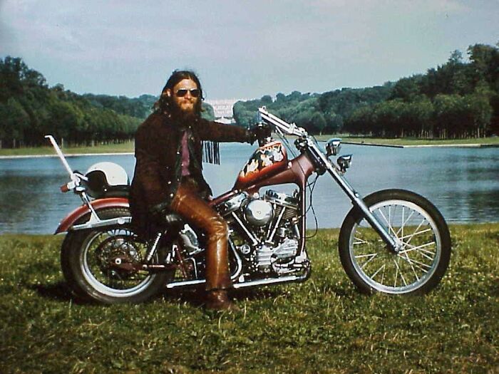 Man with sunglasses on a vintage motorcycle by a lake, capturing 1973 world in pictures vibe.