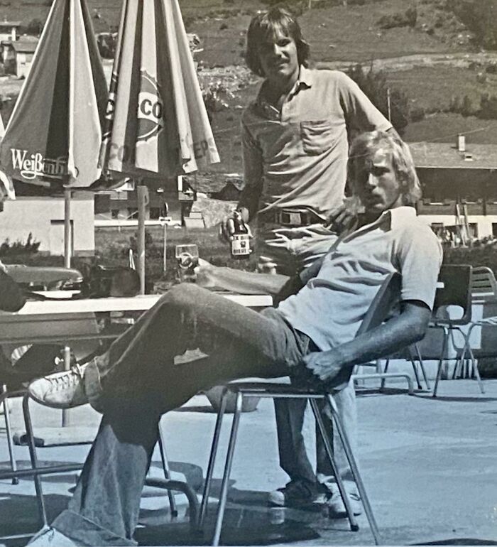 Two men relaxing outdoors with drinks in a 1973 setting, featuring classic patio umbrellas and mountainous backdrop.
