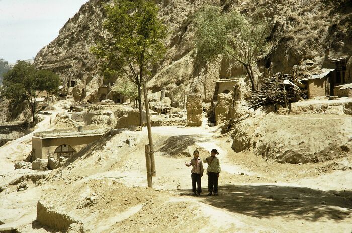 Two people walking in a 1973 village landscape, with hillside homes and trees in the background, depicting world history.