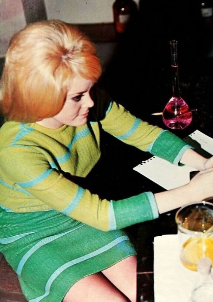 Blonde student in '70s fashion, seated at a desk with chemistry equipment.