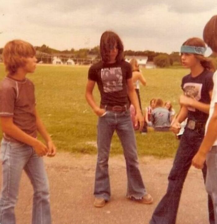 Teenagers in '70s high school fashion with flared jeans and bandanas, chatting outside on a grassy area.