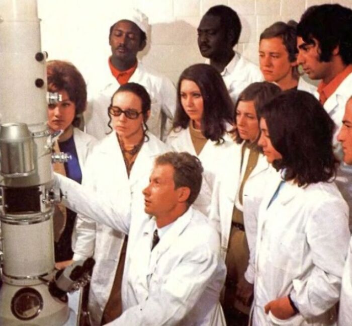 Students in the 1970s gather around a microscope in a high school science class, all wearing lab coats.