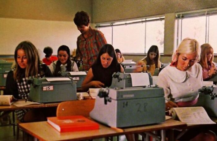 Students in a 1970s high school classroom using typewriters, focused on their work, with sunlight streaming through the windows.