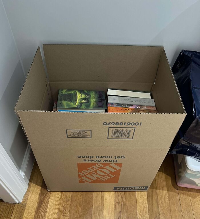 Box of books packed poorly by infuriating family members, sitting on a wooden floor.