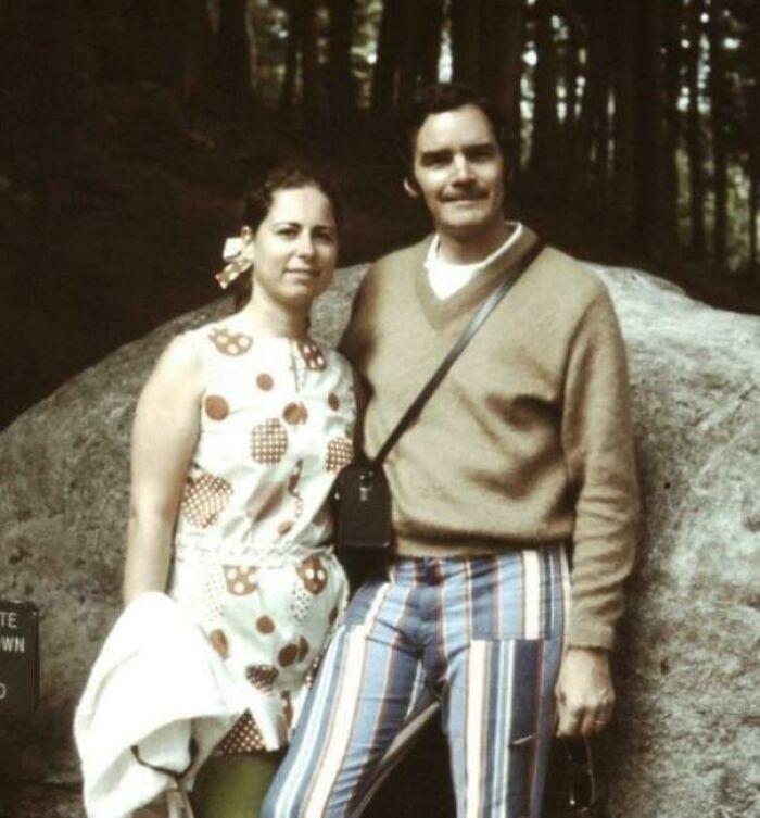 Couple in 1973 fashion, woman in polka-dot dress, man in striped pants, standing outdoors by a rock in a forest setting.