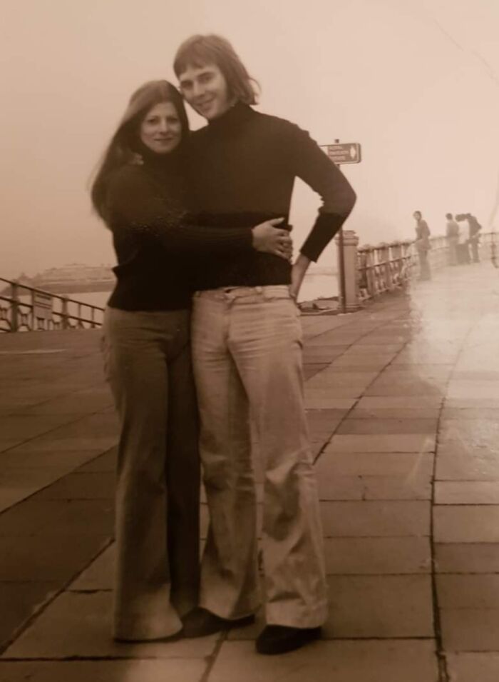 Couple in 1973, wearing turtlenecks and flared pants, on a promenade with a misty backdrop.