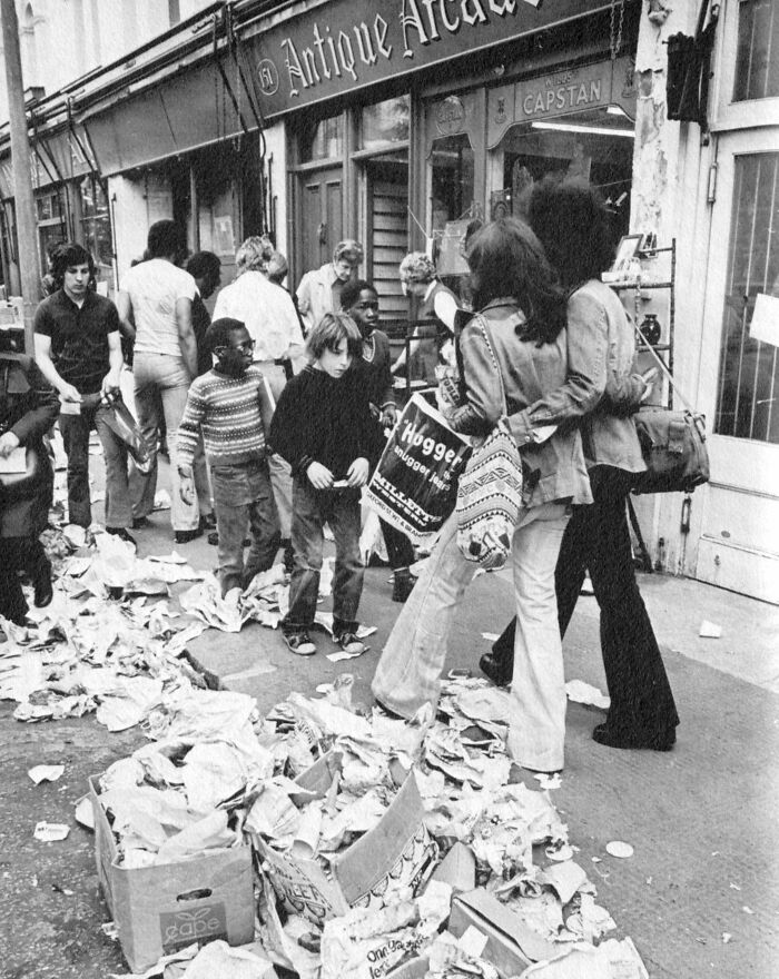 People shopping outside an antique store in 1973, surrounded by litter and holding bags in a busy street scene.