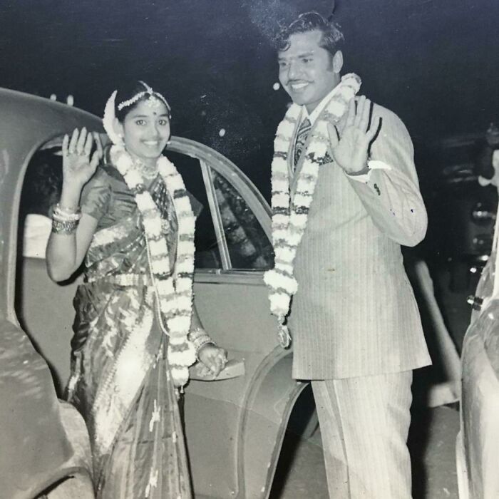 A smiling couple in wedding attire waves in front of a vintage car, showcasing a 1973 world moment.