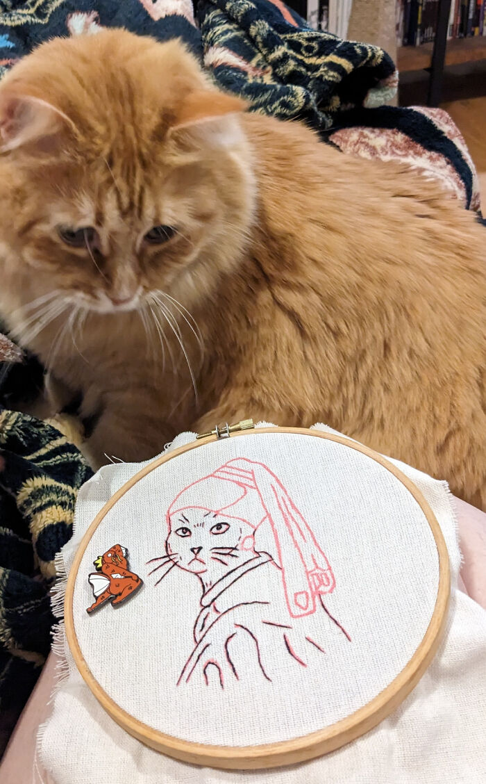 Orange cat next to an embroidery hoop featuring a cat design, showcasing beautiful embroidery designs in progress.