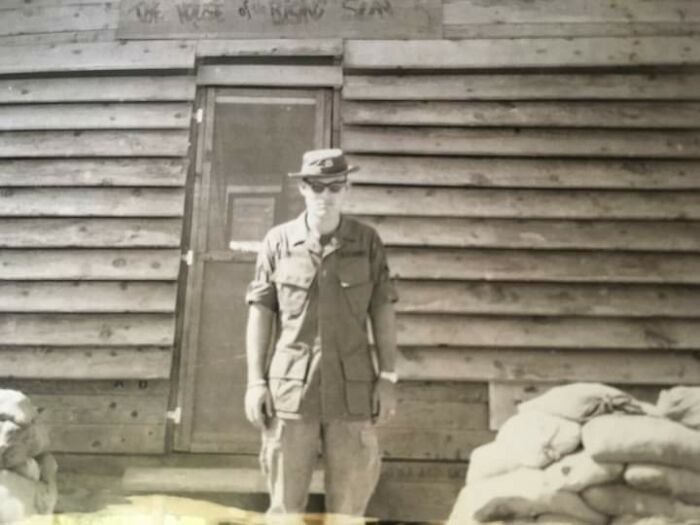 A person in military attire stands in front of a wooden building with sandbags, 1973.