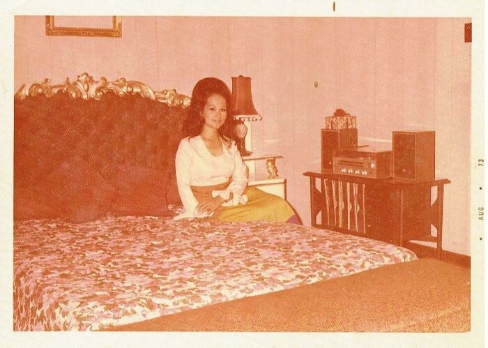 Woman sitting on a vintage bed in a 1973 room, with a retro radio and patterned bedspread, capturing a moment from the 1973 world.