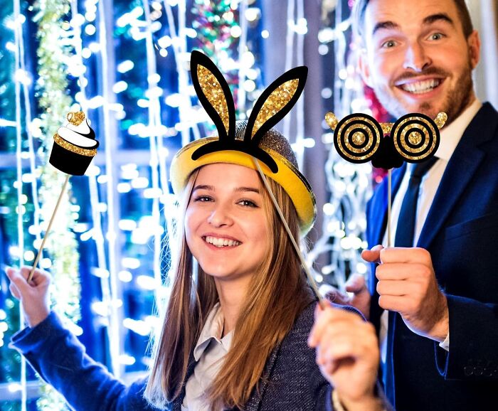 Smiling people in festive attire celebrating, using photo props, with the scene lit by colorful lights.