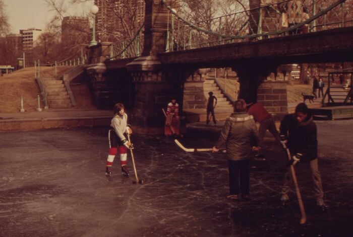 People playing hockey on a frozen pond under a bridge, reminiscent of 1973 world scenes.