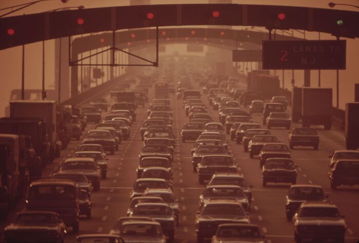 Heavy traffic on a 1973 highway during sunset, with cars and trucks moving slowly under a sign indicating lanes to New Jersey.