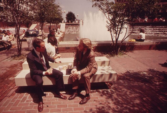Two men in suits talking on a bench in a 1973 urban park, near a fountain, with other people relaxing nearby.