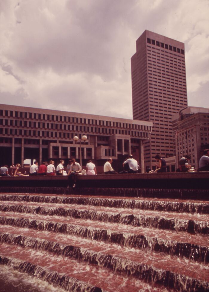 People sitting by cascading fountains in a 1973 urban setting, with tall buildings in the background under a cloudy sky.