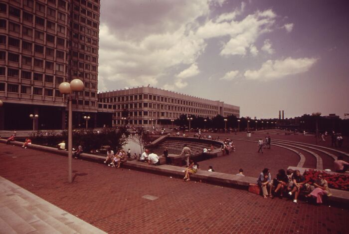 1973 urban scene with people relaxing in a city plaza, surrounded by modern buildings under a partly cloudy sky.