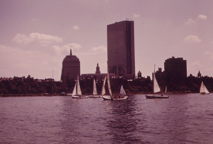 Sailboats on a river with a 1973 skyline backdrop, featuring tall buildings under a clear sky.