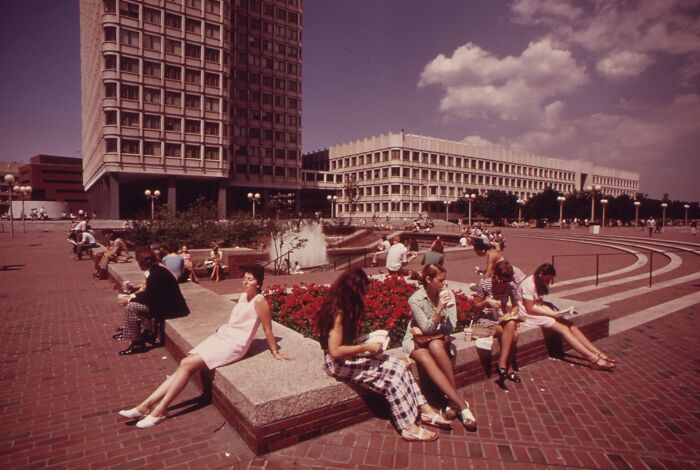 People relaxing in a plaza during 1973, surrounded by iconic buildings and a bright sky.