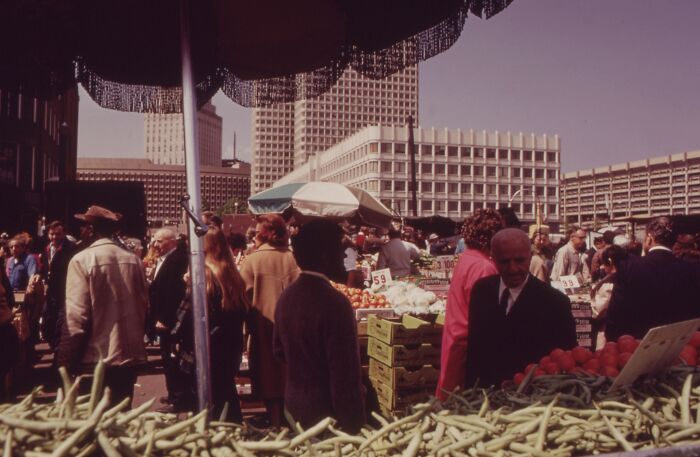1973 world market scene with bustling crowd, vegetables, and high-rise buildings in the background.