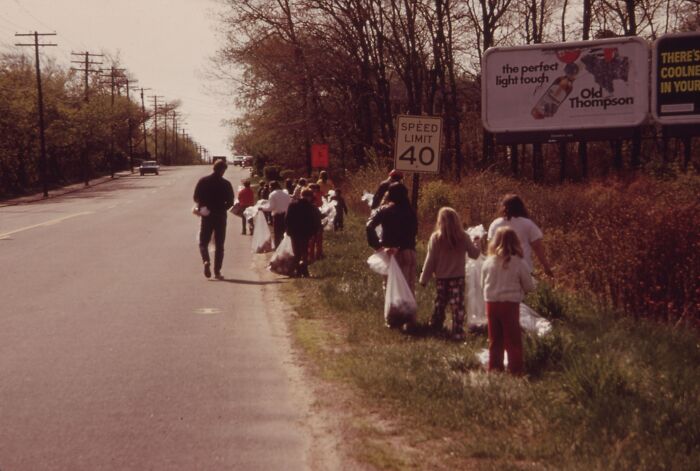 People walking on a roadside in 1973, holding bags for collecting trash, with trees and a billboard in the background.