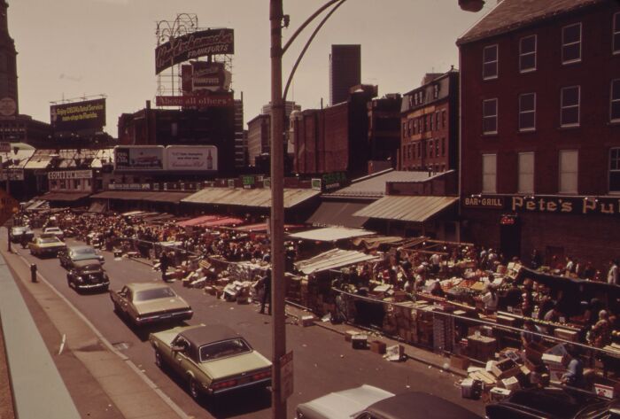 Busy street market scene, 1973, with vintage cars and bustling vendors under large advertising signs.
