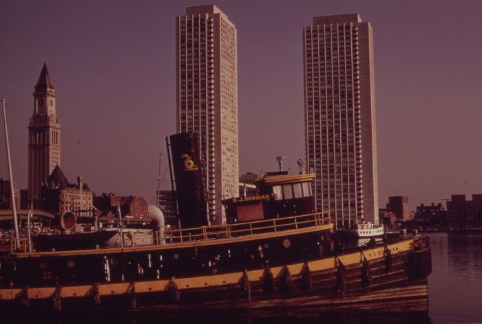 Historic tugboat in front of skyscrapers with clock tower in 1973, reflecting the world in pictures.