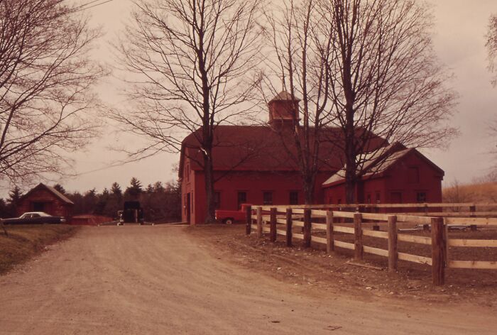 Rustic red barn and dirt road in rural 1973 setting, surrounded by bare trees, with vintage vehicles in the distance.