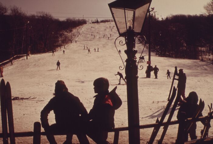 Two people silhouetted against a snowy hillside with skiers, capturing a winter scene from 1973.