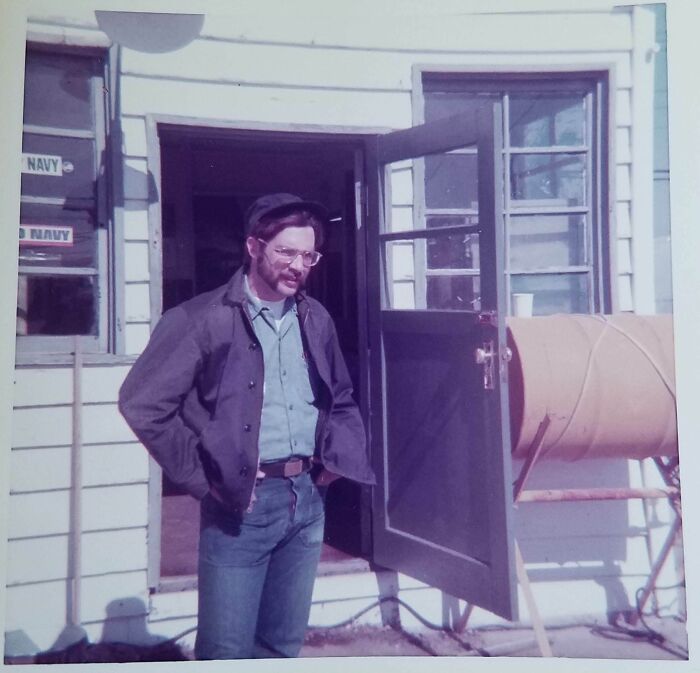 Man in 1973 standing outside a building, wearing glasses and a casual outfit, with a door open behind him.