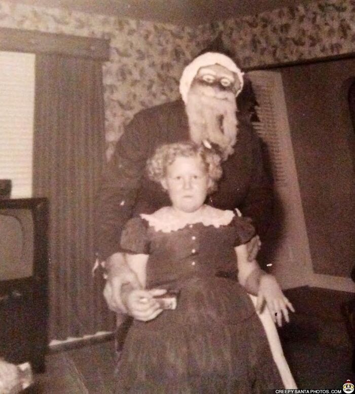 A creepy Santa with a young girl, both in vintage attire, inside a dimly lit room.