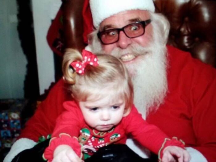 A creepy Santa with a wide grin posing with an unamused child in festive attire on his lap.