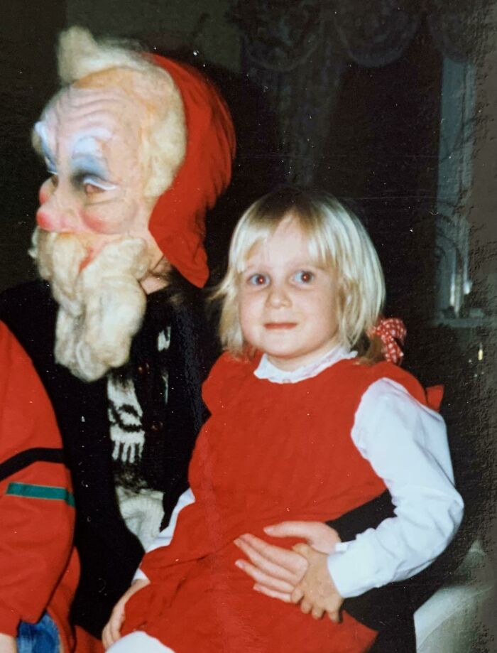 Child sitting on a person's lap dressed as creepy Santa with a red hood and mask.