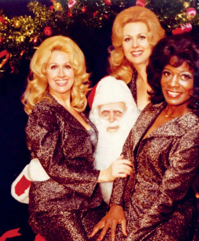 Creepy Santa surrounded by three women in sparkling outfits, posing joyfully with Christmas decorations in the background.