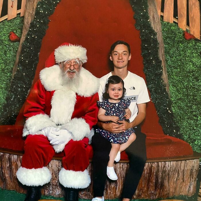 Man dressed as creepy Santa with a young girl and her father sitting on a holiday-themed backdrop.