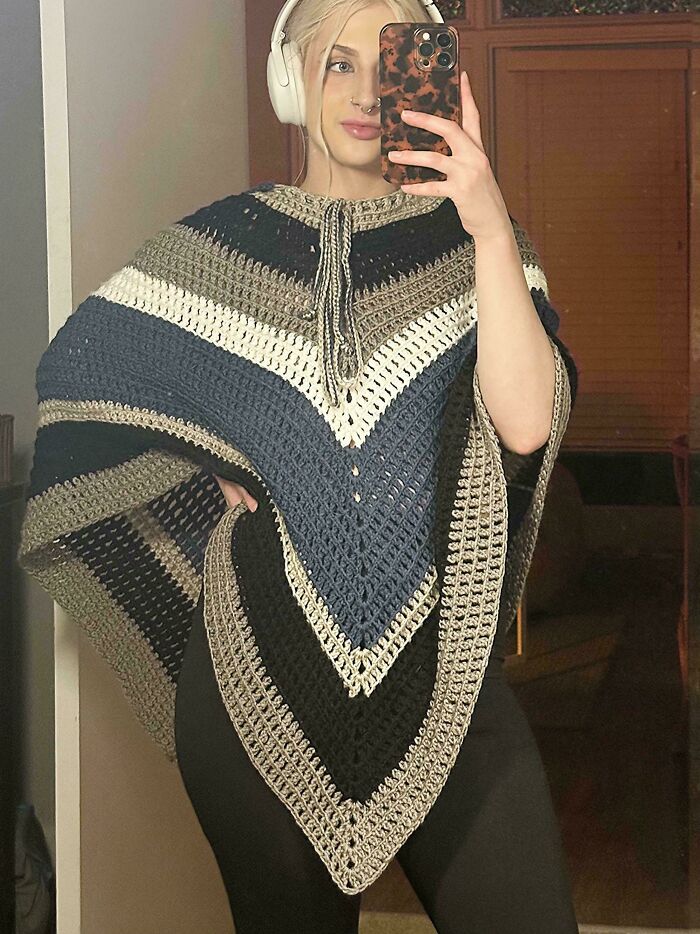 Young woman wearing headphones and a colorful handmade crochet poncho, proudly showing her crochet project.